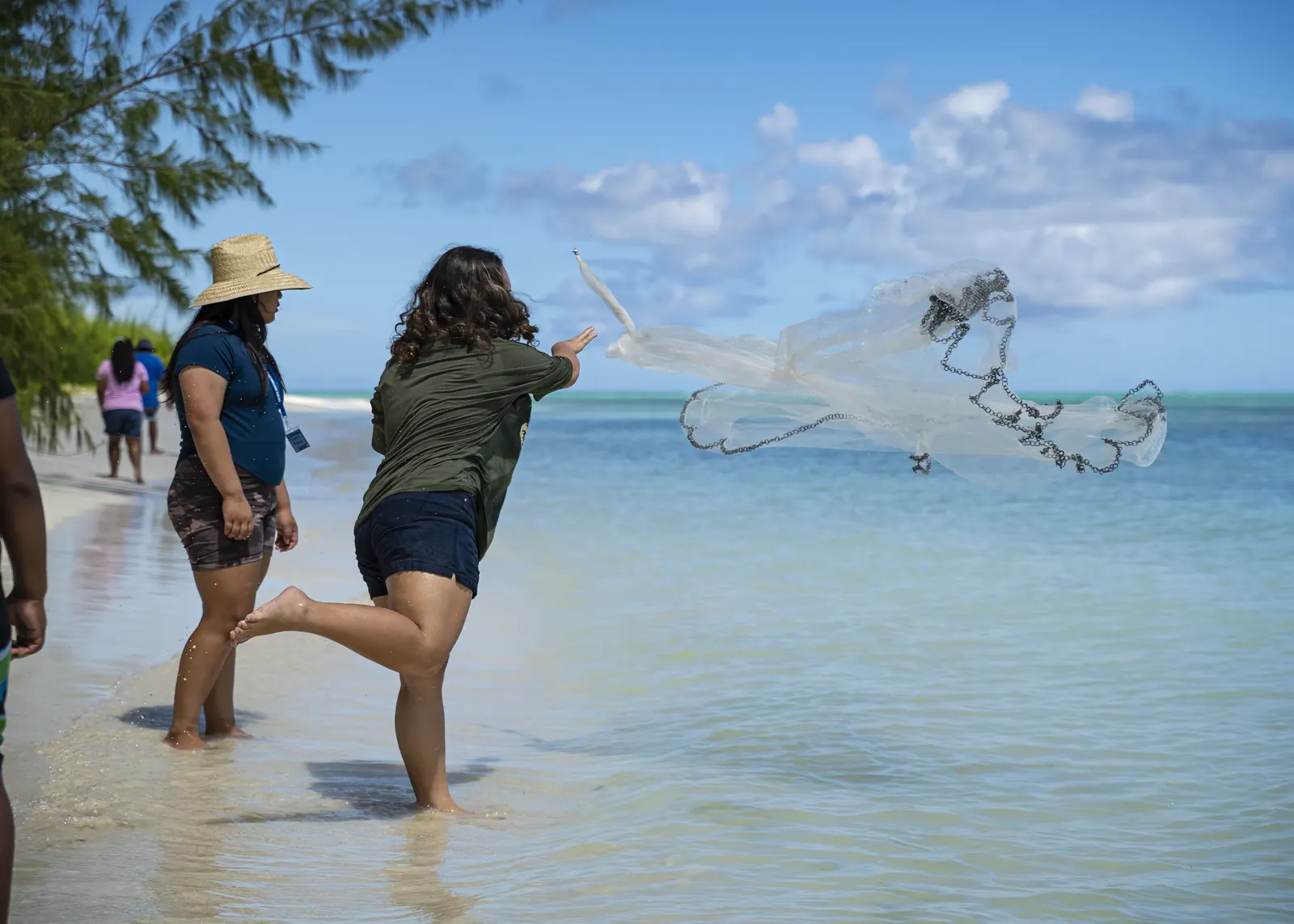Two people in the shallows, one casting a talåya net that opens in the air