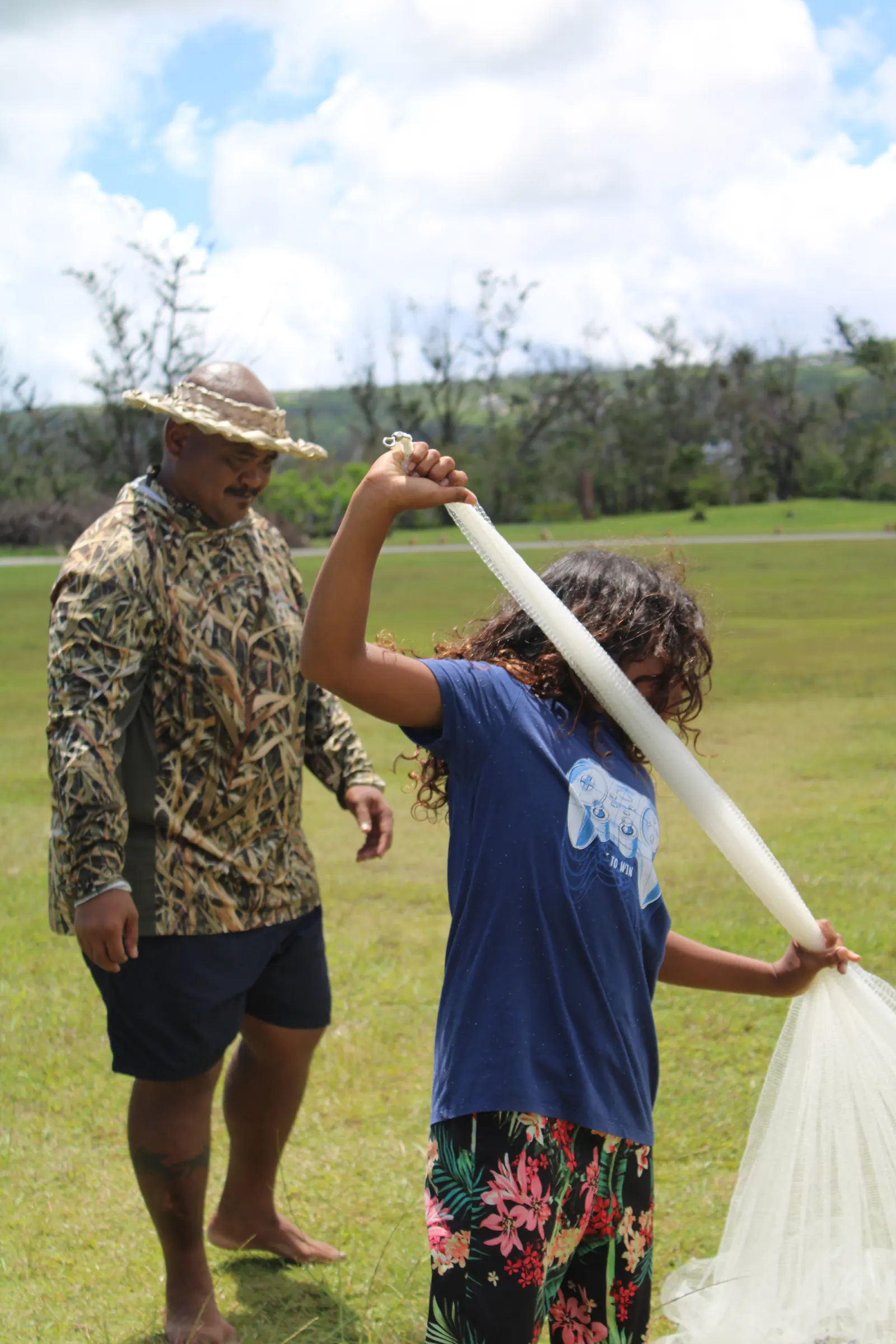 An instructor in a hat beside a young person lifting a long talåya net on a grassy field